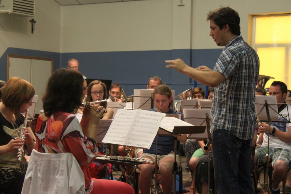 Thomas Groenweghe dirige l'orchestre dans l'ancienne salle des fêtes de Montzen. Les musiciens sont habillés en civil.